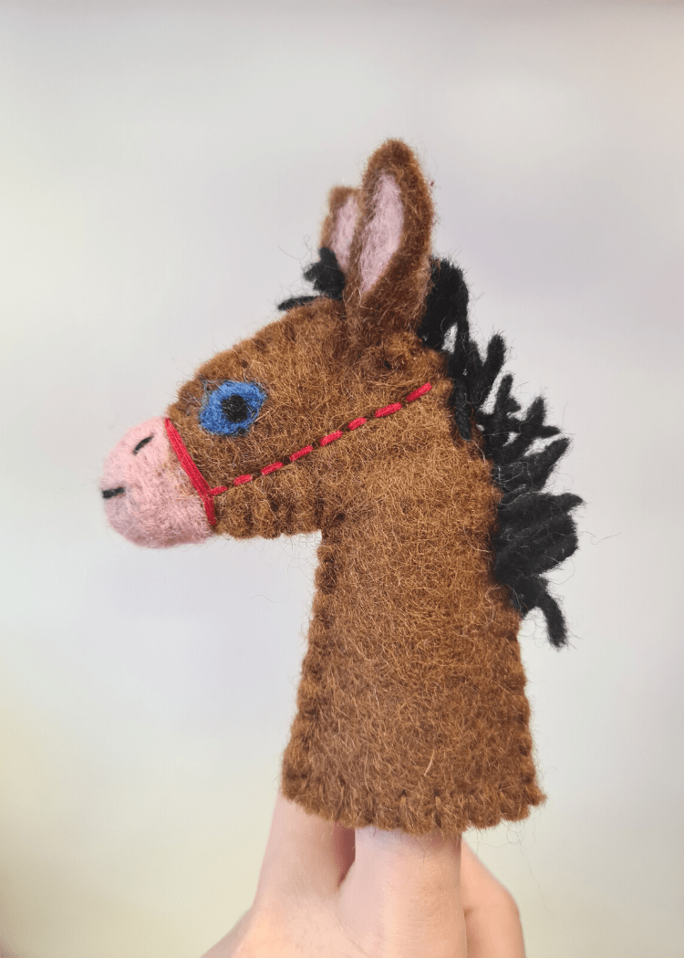 brown felt horse finger puppet with a black main, pink nose, blue eye and red bridle sat on a persons finger