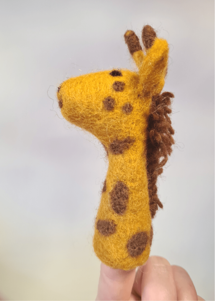 side view of a yellow felt giraffe finger puppet with brown spots and mane sat on someones finger