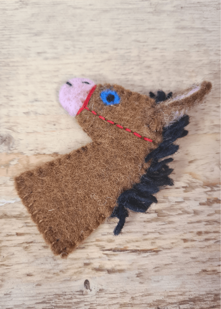 brown felt horse finger puppet with a black main, pink nose, blue eye and red bridle laid on a wooden surface