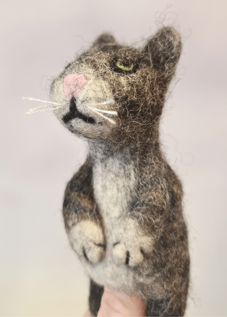 close up of grey stripy felt cat finger puppet with white whiskers and pink nose sat on someones finger