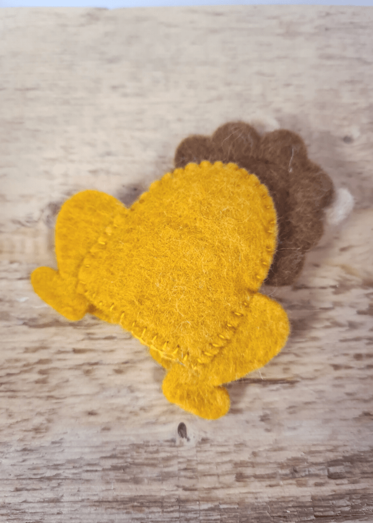 back of a sitting felt yellow lion finger puppet with brown mane laying on a wooden surface