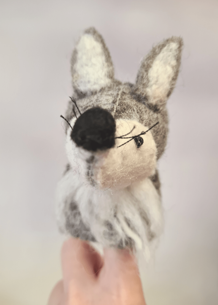 close up of a felt grey and white wolf finger puppet with black nose, whiskers and bead eyes sat on someone&