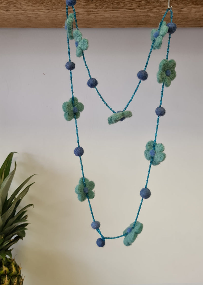 Blue pom pom flower necklace with beads hanging from a block of wood next to a pineapple