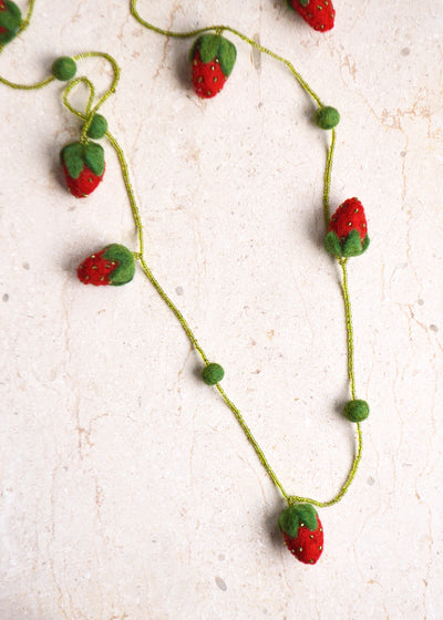 red and green felt strawberry necklace with beads and pompoms laying on a marble surface