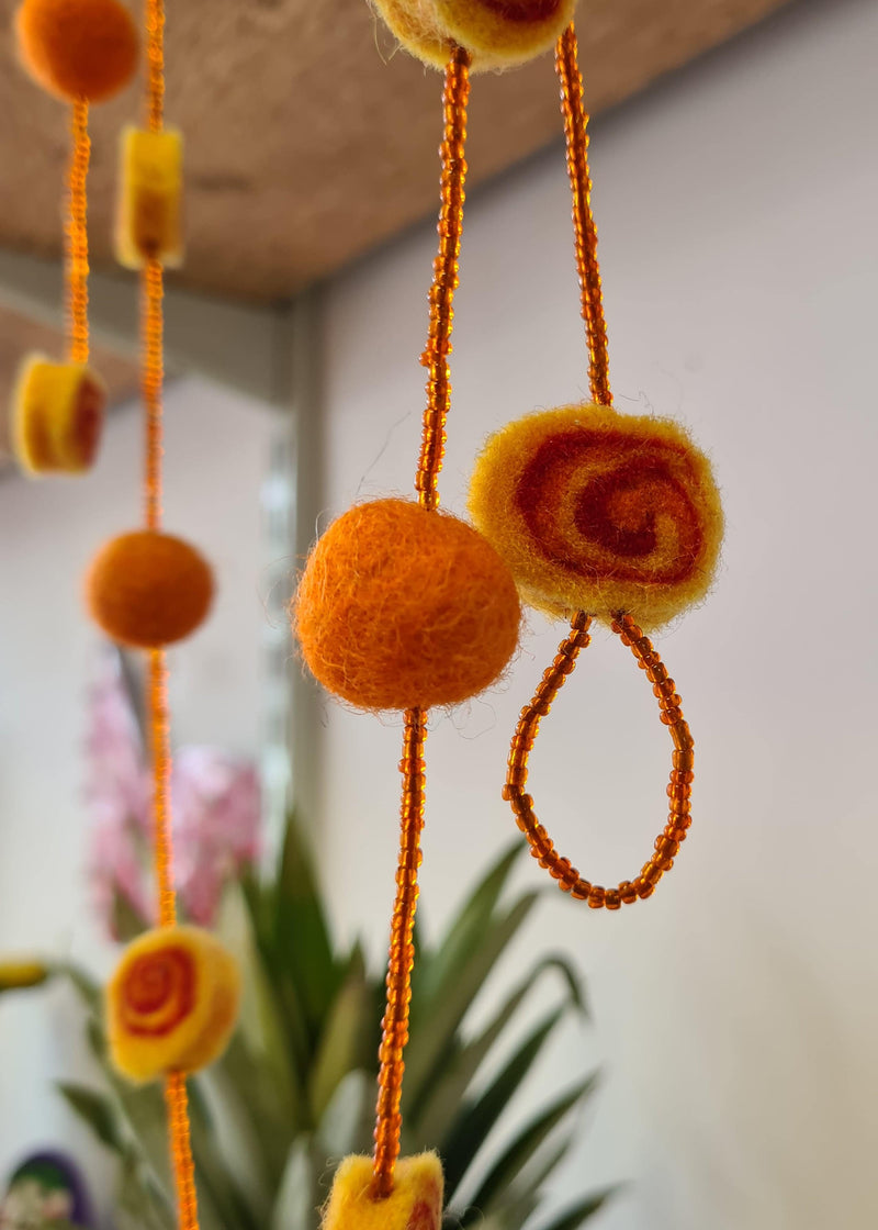 close up of Orange Felt Pom Pom Necklace With Beads and swirl detail hanging in front of a pineapple