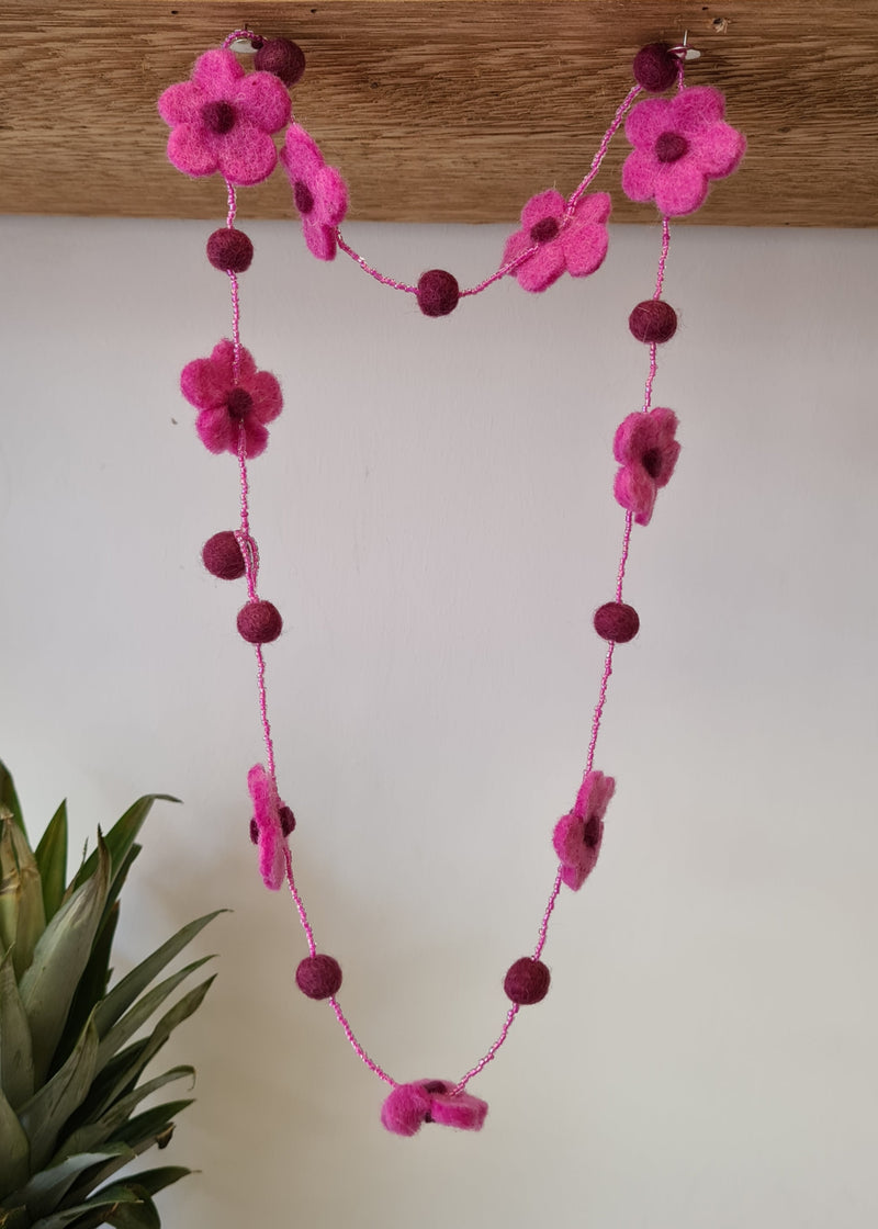 Pink pom pom flower necklace with beads hanging from a block of wood next to the top of a pineapple