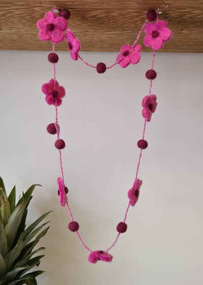 Pink pom pom flower necklace with beads hanging from a block of wood next to the top of a pineapple