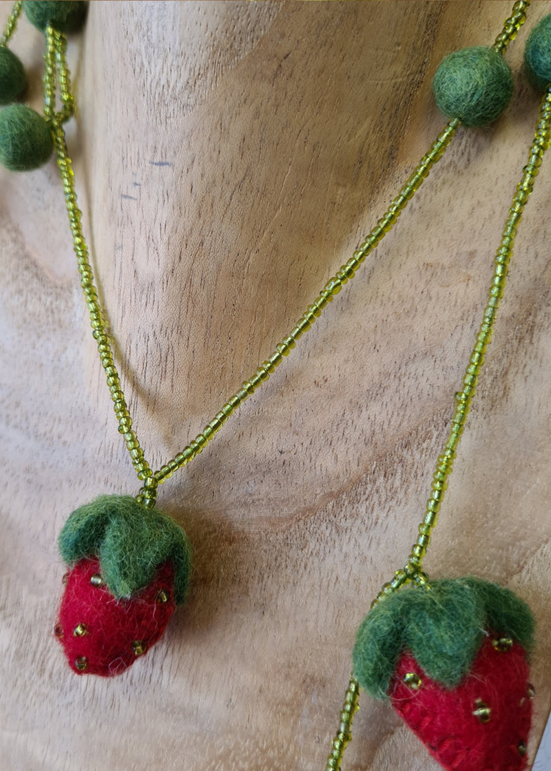 close up of strawberries and beads from a red and green felt strawberry necklace with beads and pompoms hanging on a wooden mannequin
