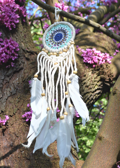 a white dreamcatcher with different shades of blue crocheted in the centre and white threads with feathers hanging from the bottom with wooden beads, hung on a tree with purple flowers in the background