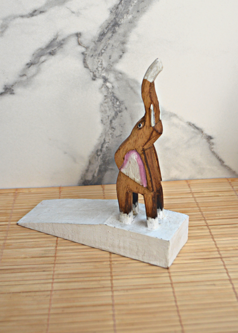 side view of a wooden brown and white elephant door stop with pink detail on ears, stood on a bamboo matt with marble background