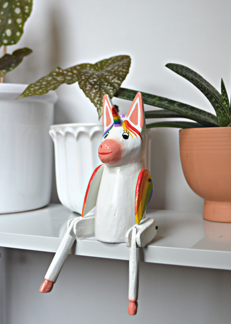 white and pink wooden unicorn with rainbow horn and wings sat on a white wooden shelf with plants in pots in the background