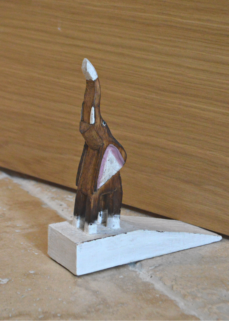 side view of a wooden brown and white elephant door stop with pink detail on ears, stood on a stone floor holding open a brown door