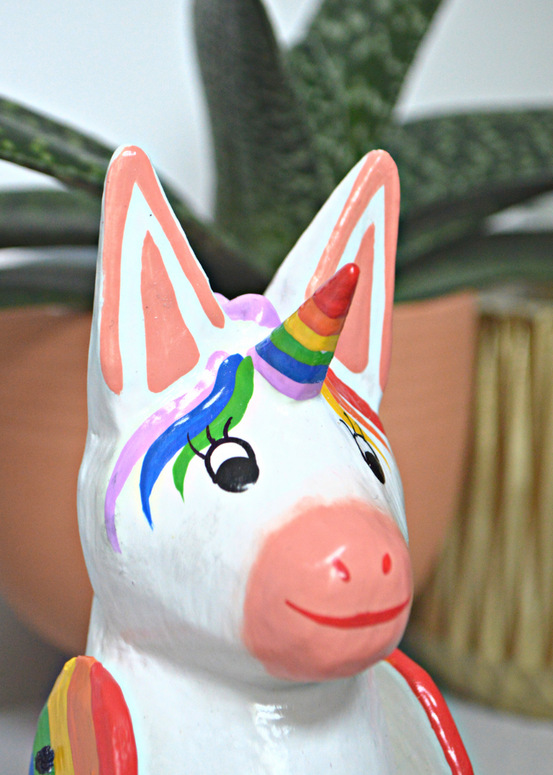 close up of white and pink wooden unicorn with rainbow horn with plants in pots in the background