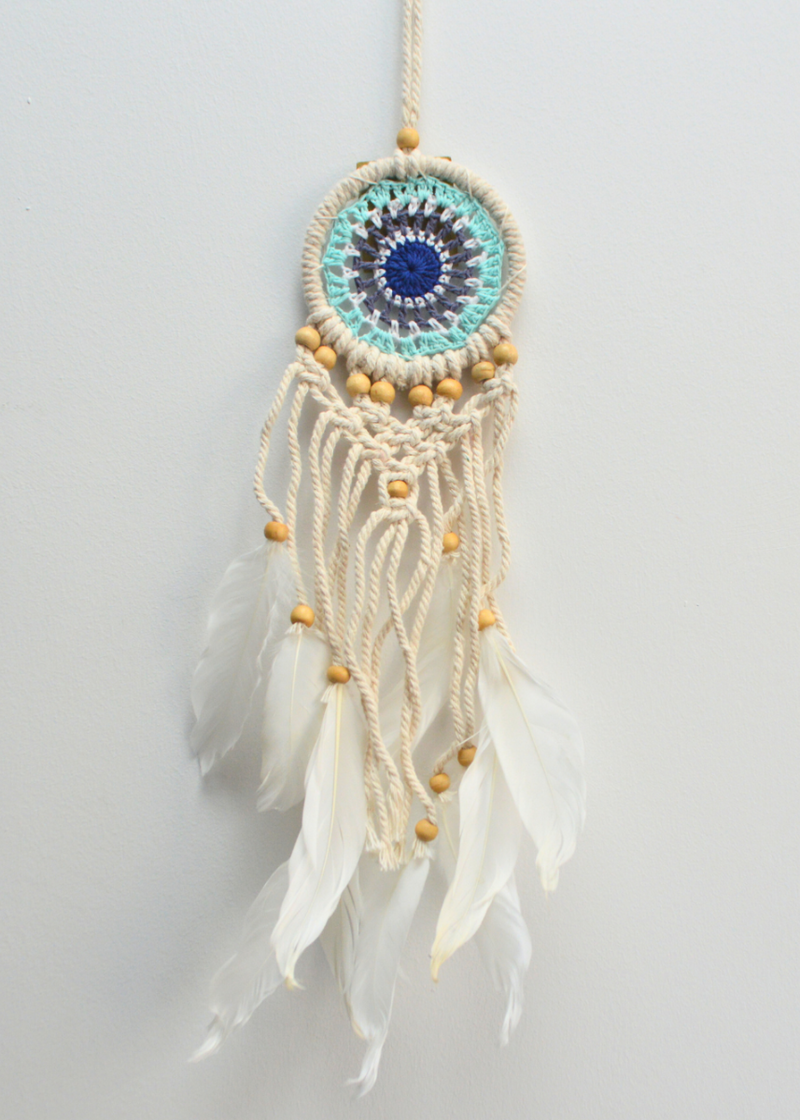 a white dreamcatcher with different shades of blue crocheted in the centre and white threads with feathers hanging from the bottom with wooden beads, hung in front of a white background
