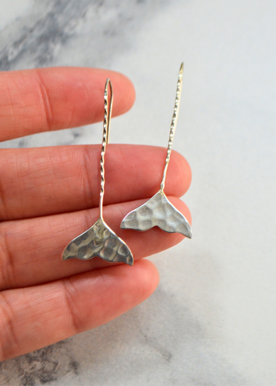 925 sterling silver whale tail earrings with long fish hook placed on a hand in front of a white marble background