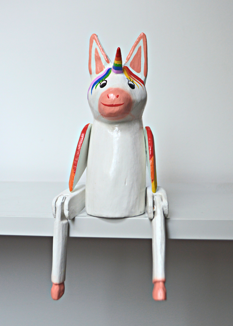 white and pink wooden unicorn with rainbow horn and wings sat on a white wooden shelf in front of white background