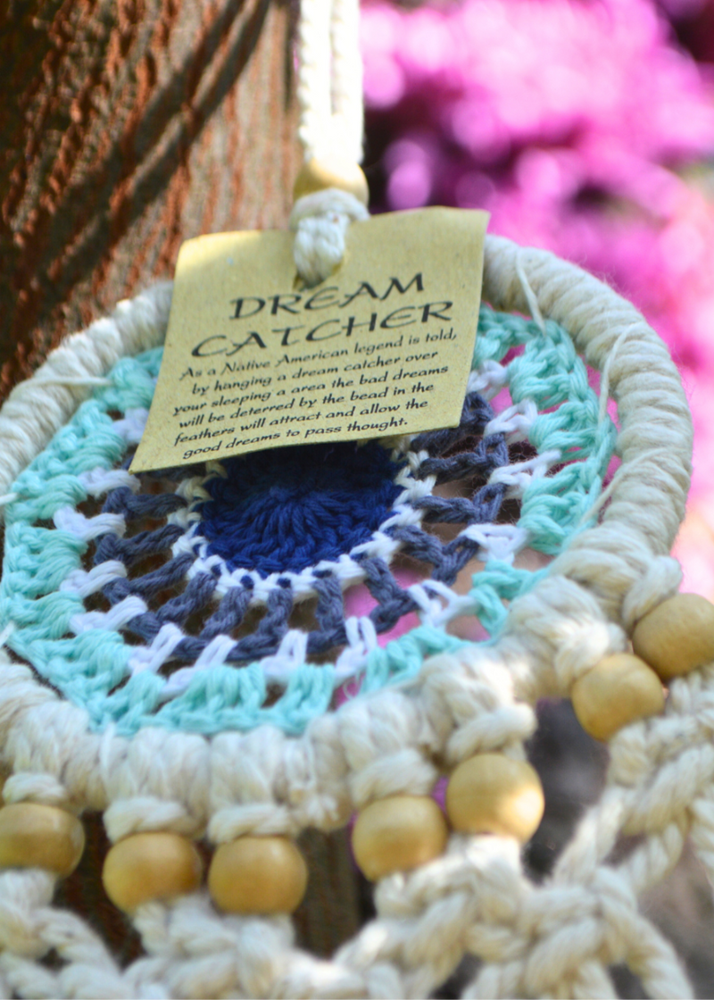 close up of a white dreamcatcher with different shades of blue crocheted in the centre and white threads hanging from the bottom with wooden beads, hung on a tree with purple flowers in the background