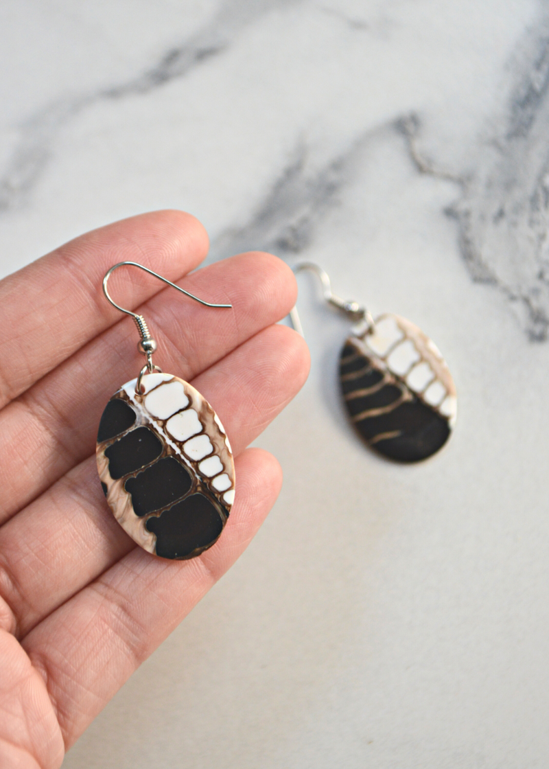 one brown and white Oval Shell Earrings With Fossil Detail and ear wire laid in a hand and the other blurred in the background on a marble surface
