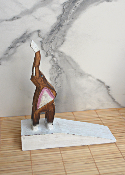 side view of a wooden brown and white elephant door stop with pink detail on ears, stood on a bamboo matt with marble background