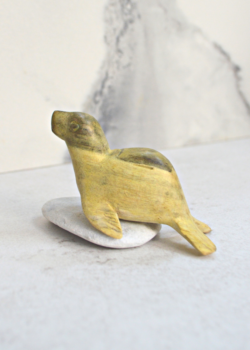 a small green and yellow wooden seal decoration sat on a pebble from a side view with a white marble background