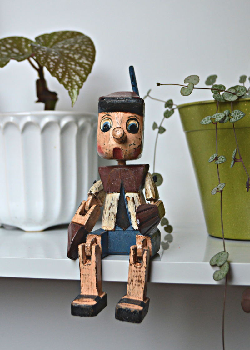 Shabby chic wooden pinnochio shelf sitter sat on a white shelf with plants in the background