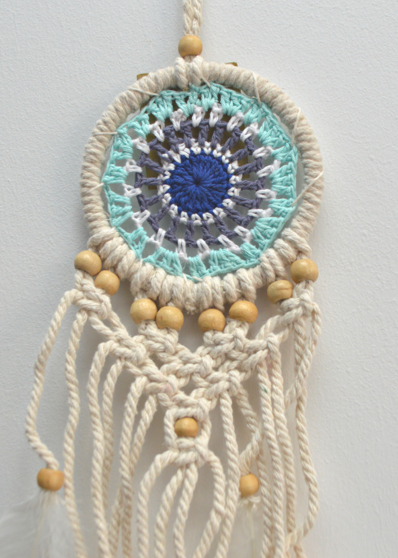a white dreamcatcher with different shades of blue crocheted in the centre and white threads hanging from the bottom with wooden beads, hung in front of a white background
