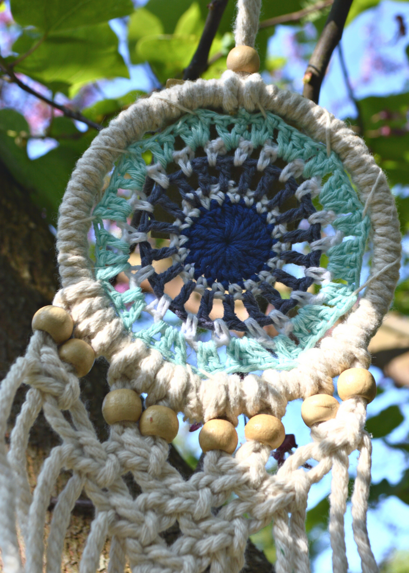 a white dreamcatcher with different shades of blue crocheted in the centre and white threads hanging from the bottom with wooden beads, hung on a tree