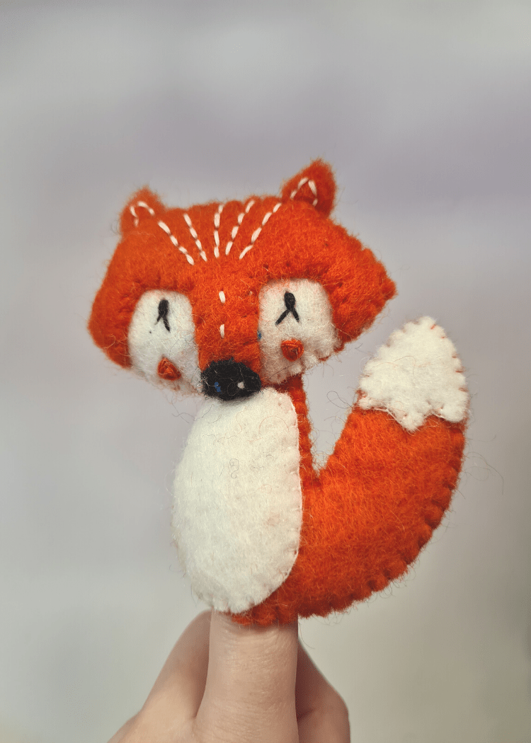 felt orange fox finger puppet with white belly eyes and tail end and black nose sat on someones finger
