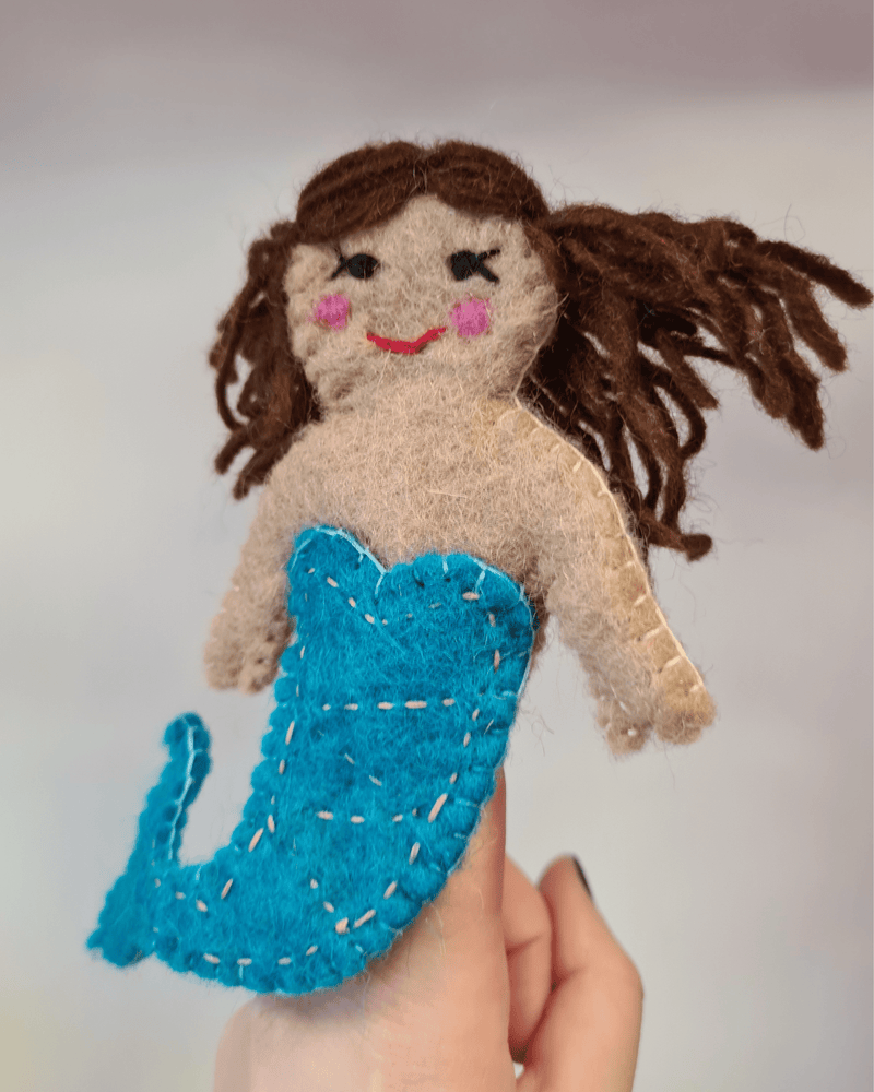 Elefair Girftware felt mermaid finger puppet with a blue tail with brown hair and rosy red cheeks sat on a finger