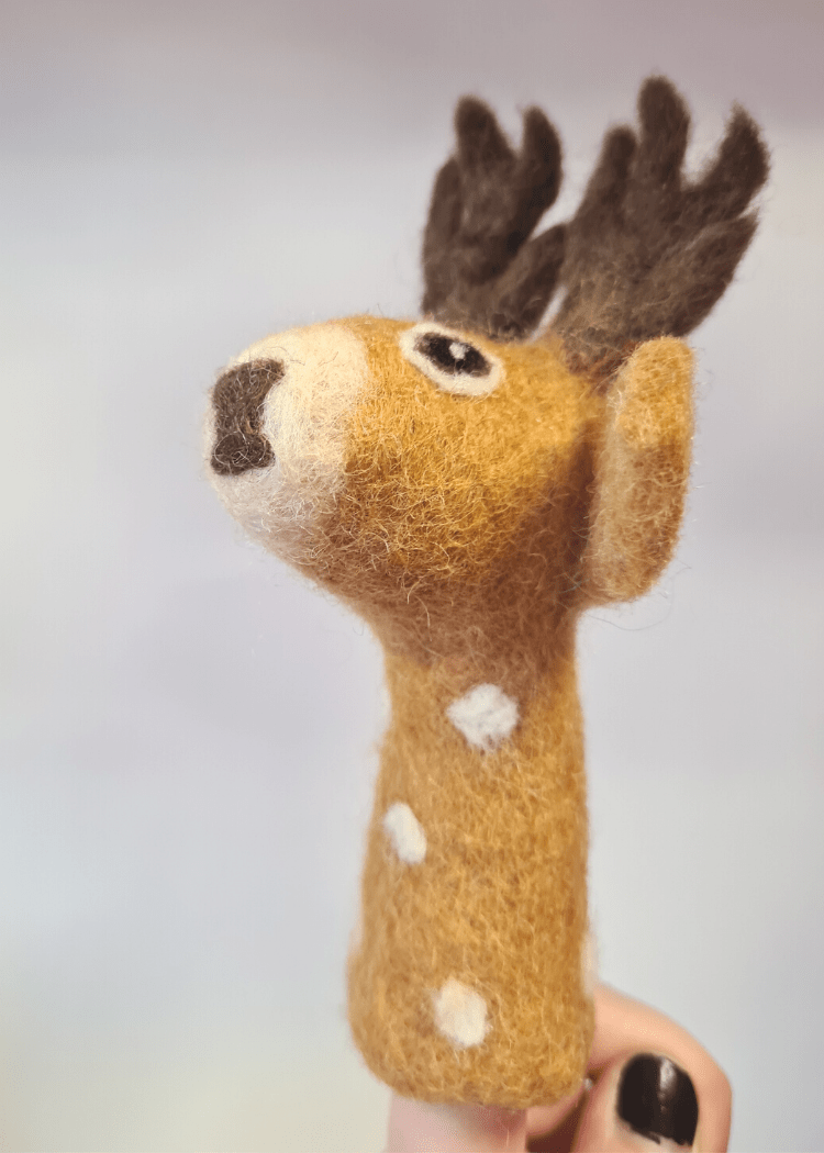 side view of a tan brown felt finger puppet with white spots and dark brown antlers and nose sat on someones finger