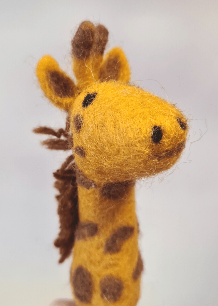 close up of a yellow felt giraffe finger puppet with brown spots and mane sat on someones finger
