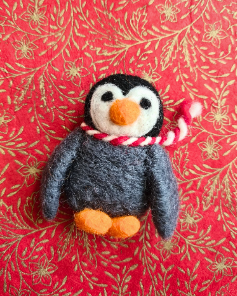 a felt penguin brooch with an orange nose, red & white scarf and grey body laid on a red and gold background
