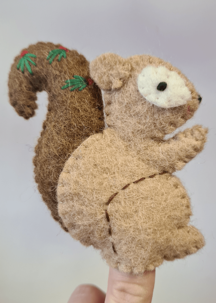close up of light brown felt squirrel finger puppet with dark brown tail and leaves on, sat on someones finger