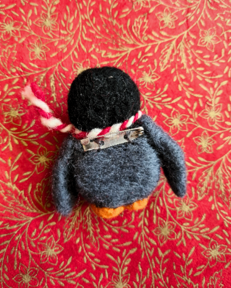 back view of a felt penguin brooch with a orange feet, red & white scarf and grey body laid on a red and gold background