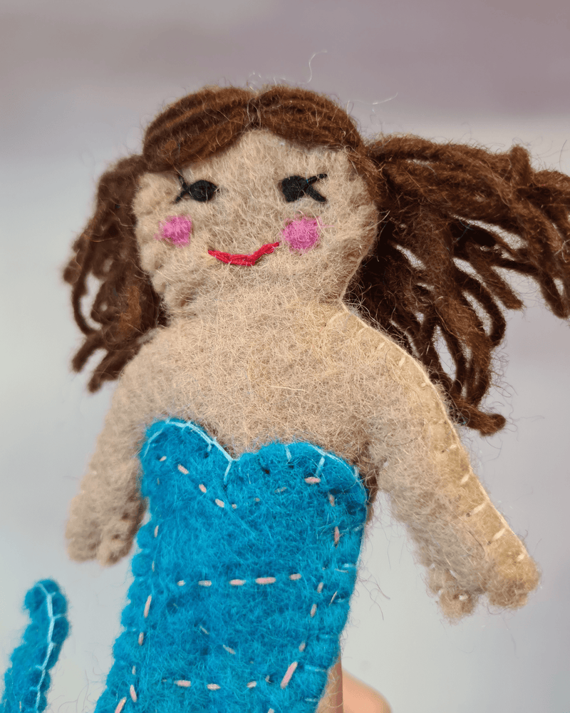 close up of Elefair Girftware felt mermaid finger puppet with a blue tail with brown hair and rosy red cheeks sat on a finger
