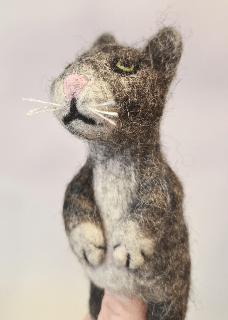 close up of grey stripy felt cat finger puppet with white whiskers and pink nose sat on someones finger 