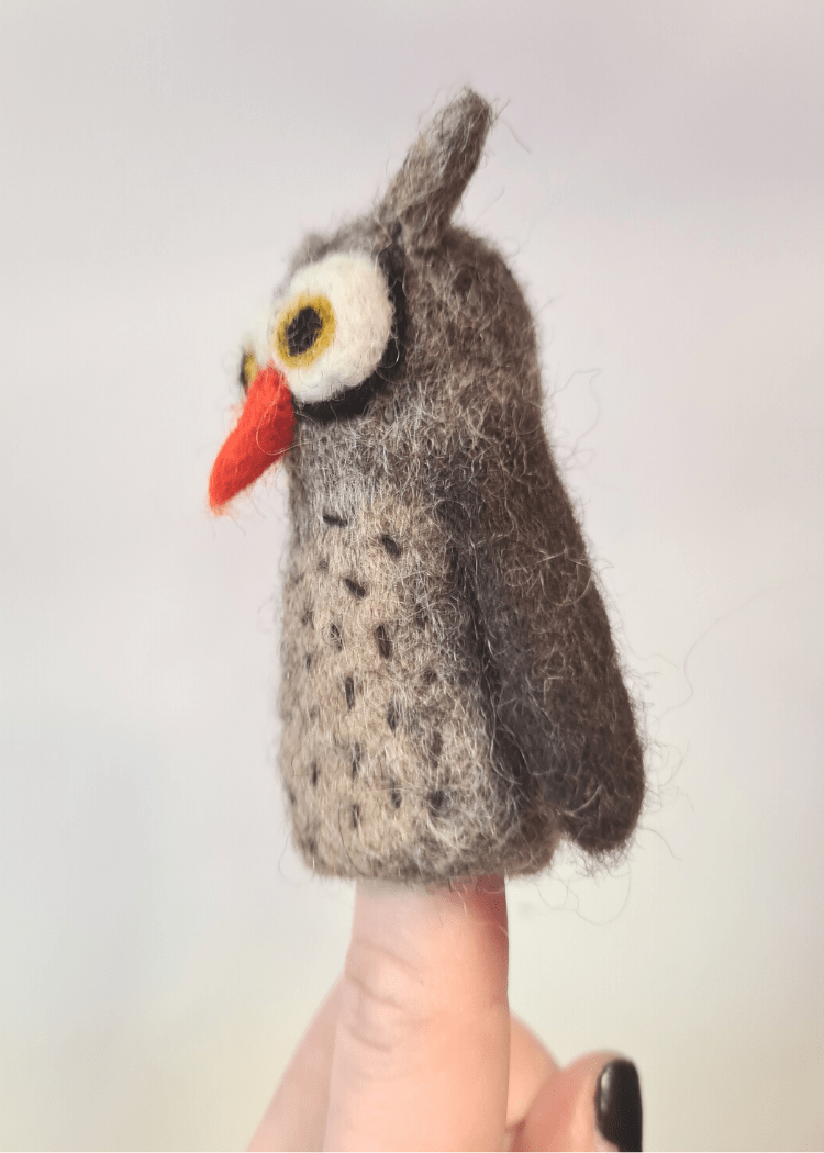side view of a brown felt owl finger puppet with orange nose and big white and brown eyes sat on someone&