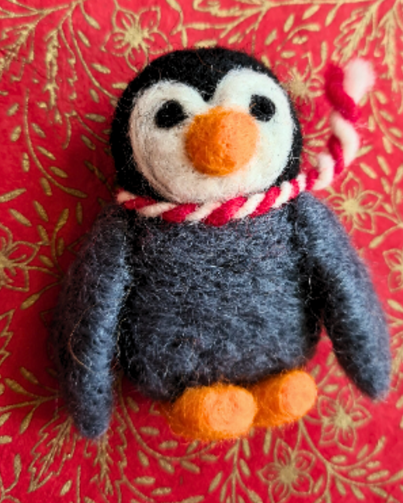 close up of a felt penguin brooch with an orange nose, red & white scarf and grey body laid on a red and gold background