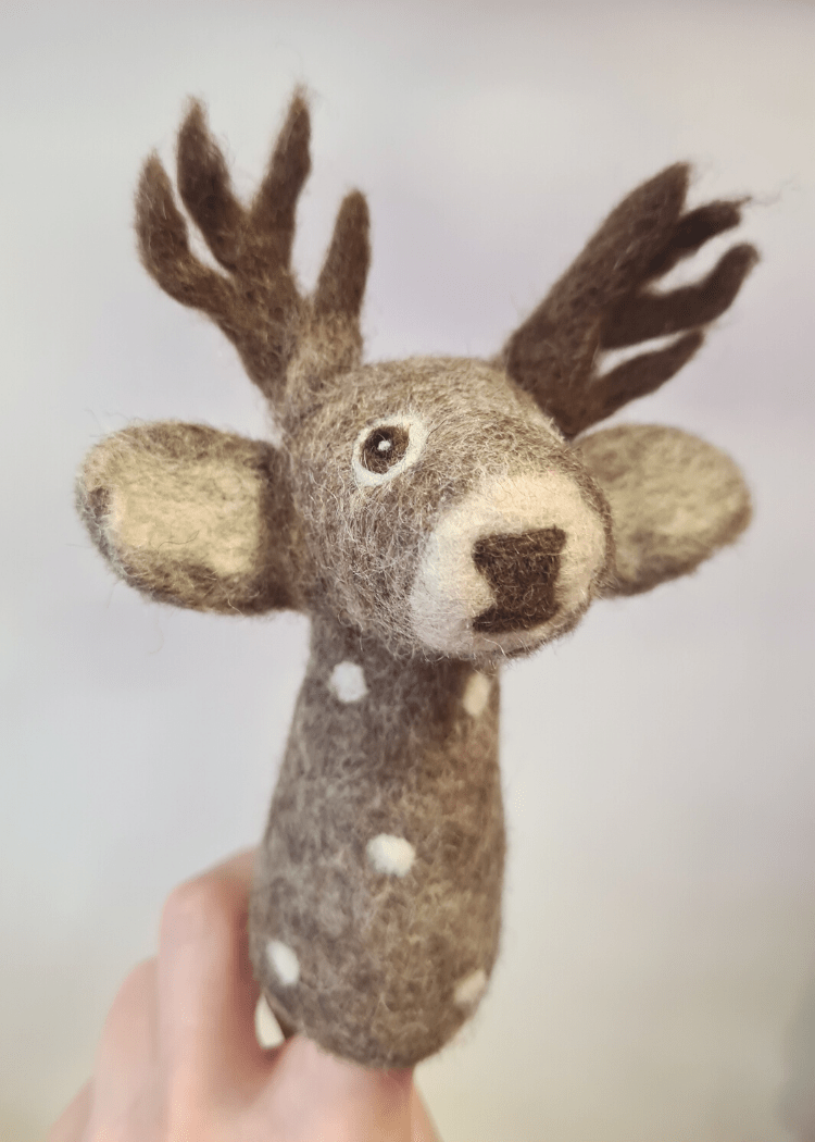 close up of a grey felt deer finger puppet with white spots, brown antlers and nose, a cute face and big ears sat on someones finger