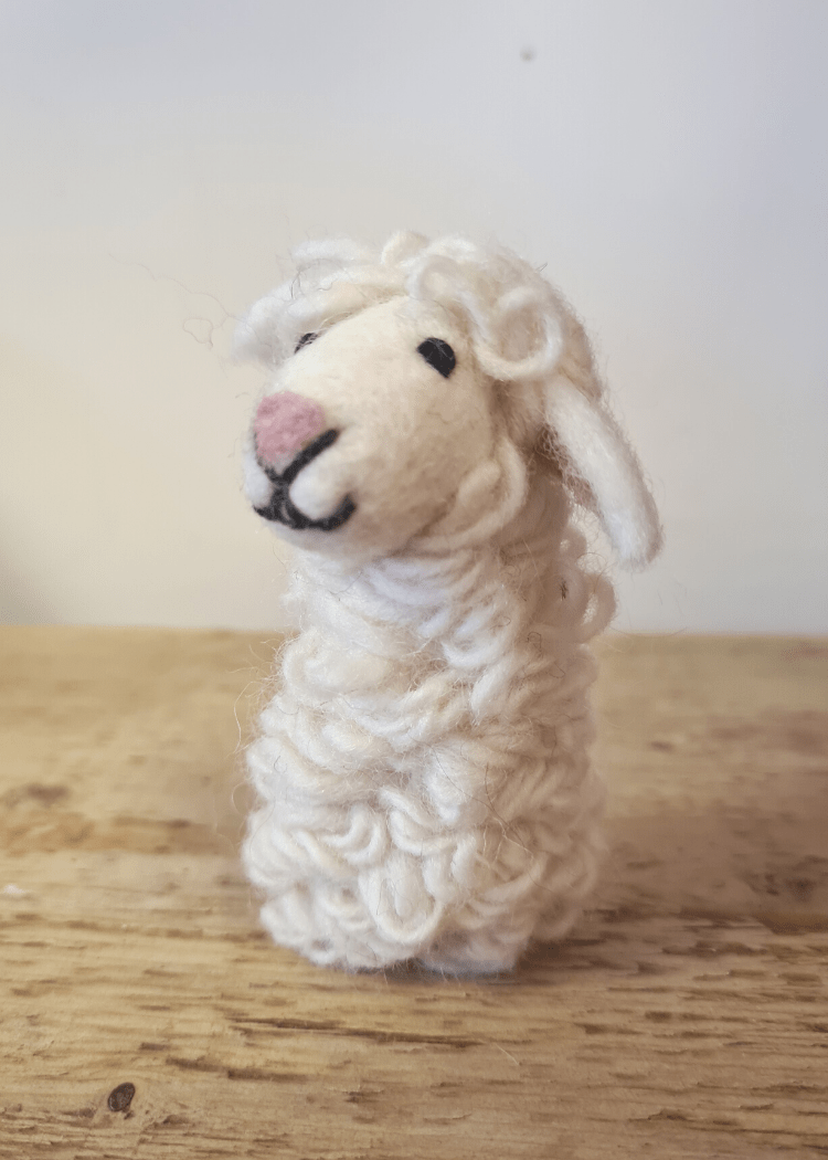 white felt sheep finger puppet with pink nose and black thread smile and eyes sat on wooden top