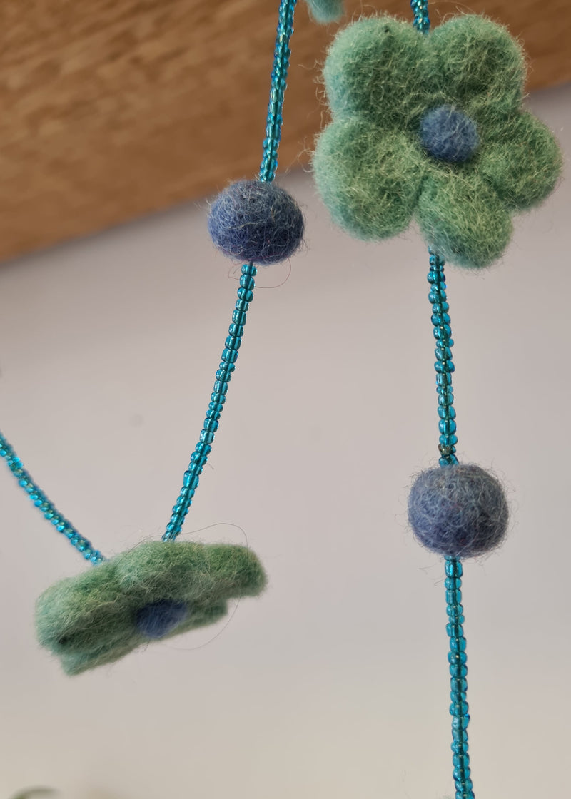 Blue pom pom flower necklace with beads hanging from wooden block with close up of flower