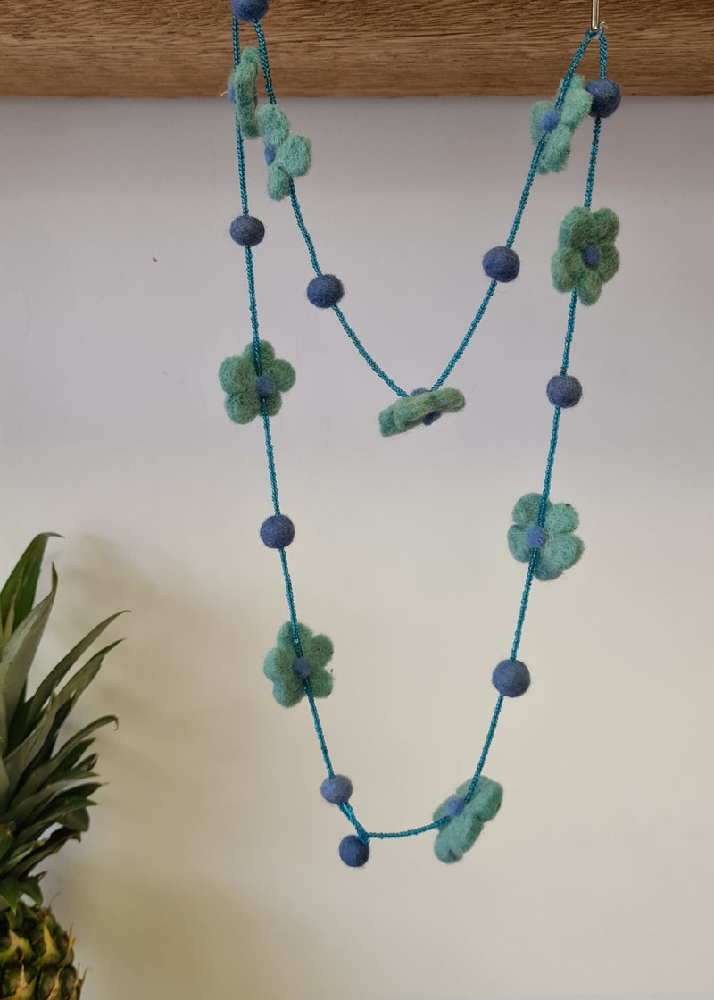 Blue pom pom flower necklace with beads hanging from a block of wood next to a pineapple 