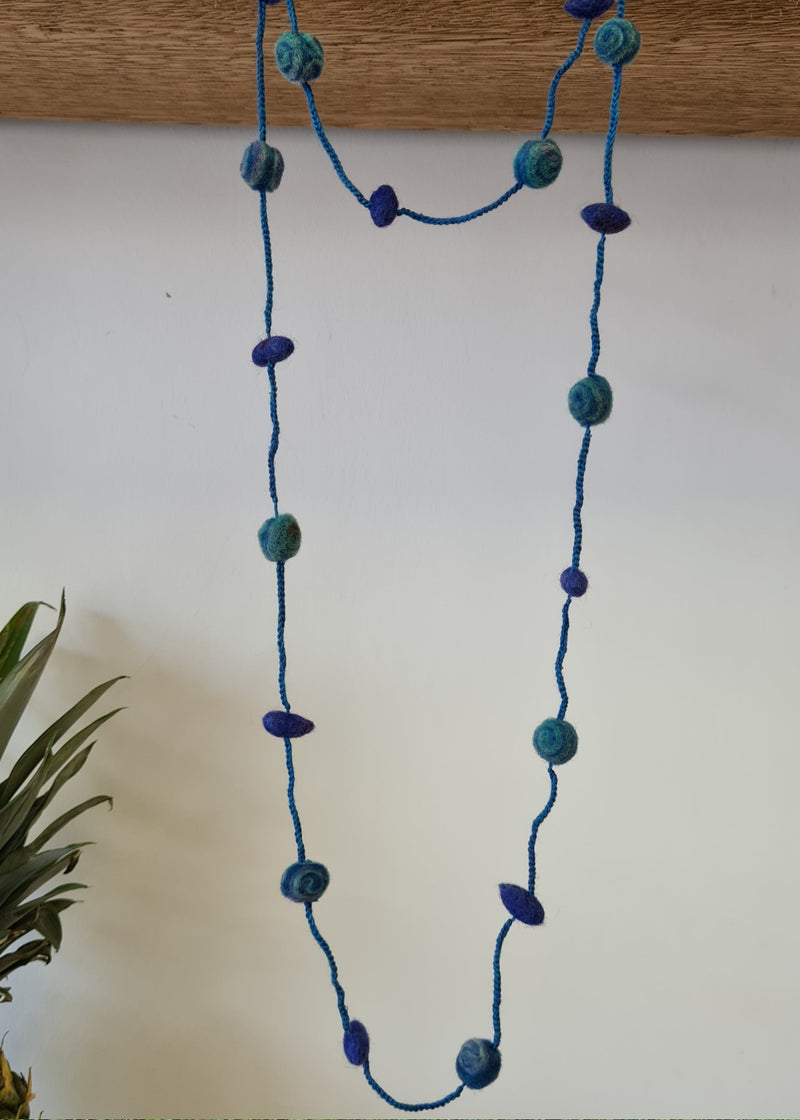 Blue Pom Pom Felt Necklace hanging from a plank of wood next to a pineapple