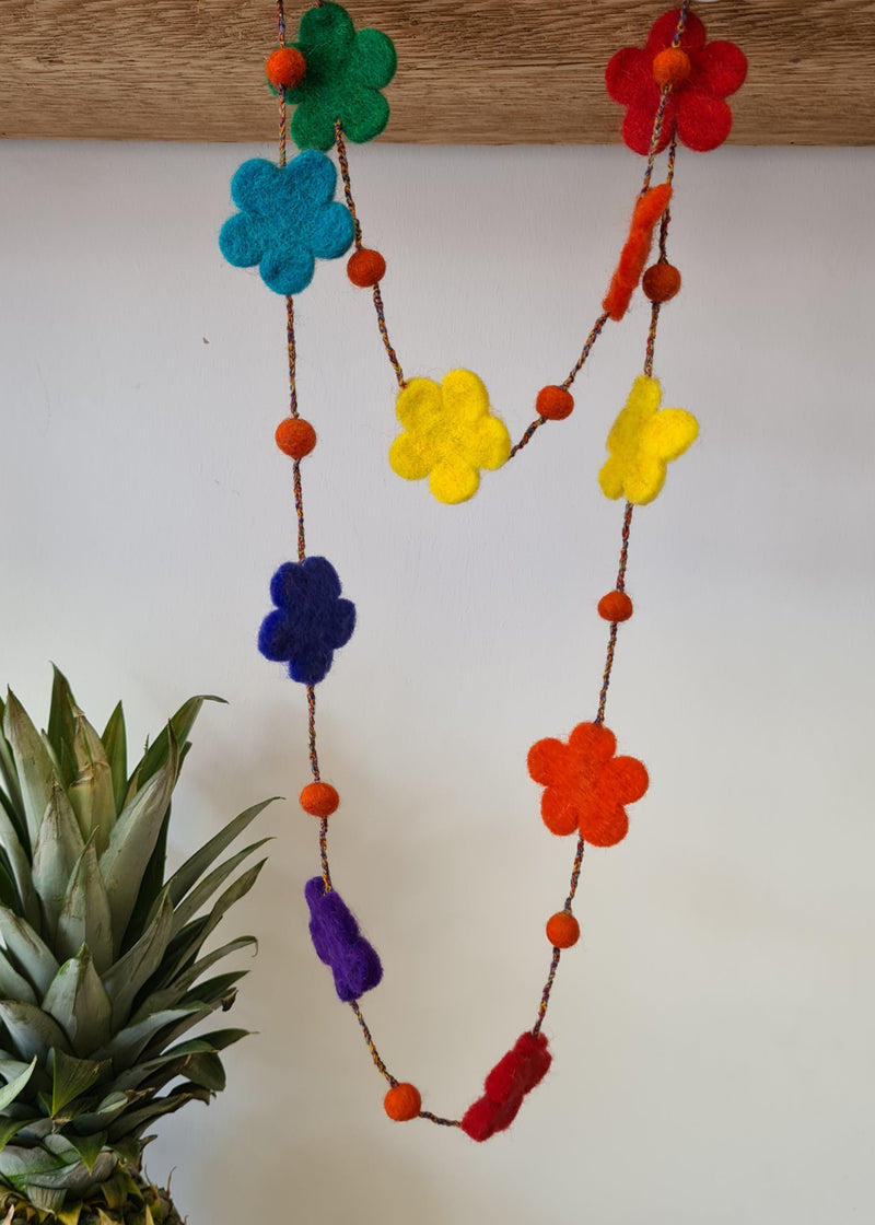 multi-colour felt pom pom flower necklace hanging from a block of wood next to a pineapple
