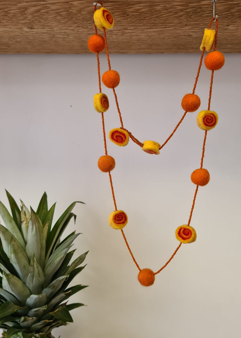 Orange  Felt Pom Pom Necklace With Beads hanging next to a pineapple