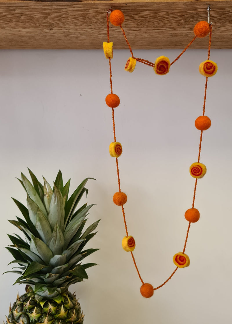 Orange Felt Pom Pom Necklace With Beads and swirl detail hanging next to a pineapple