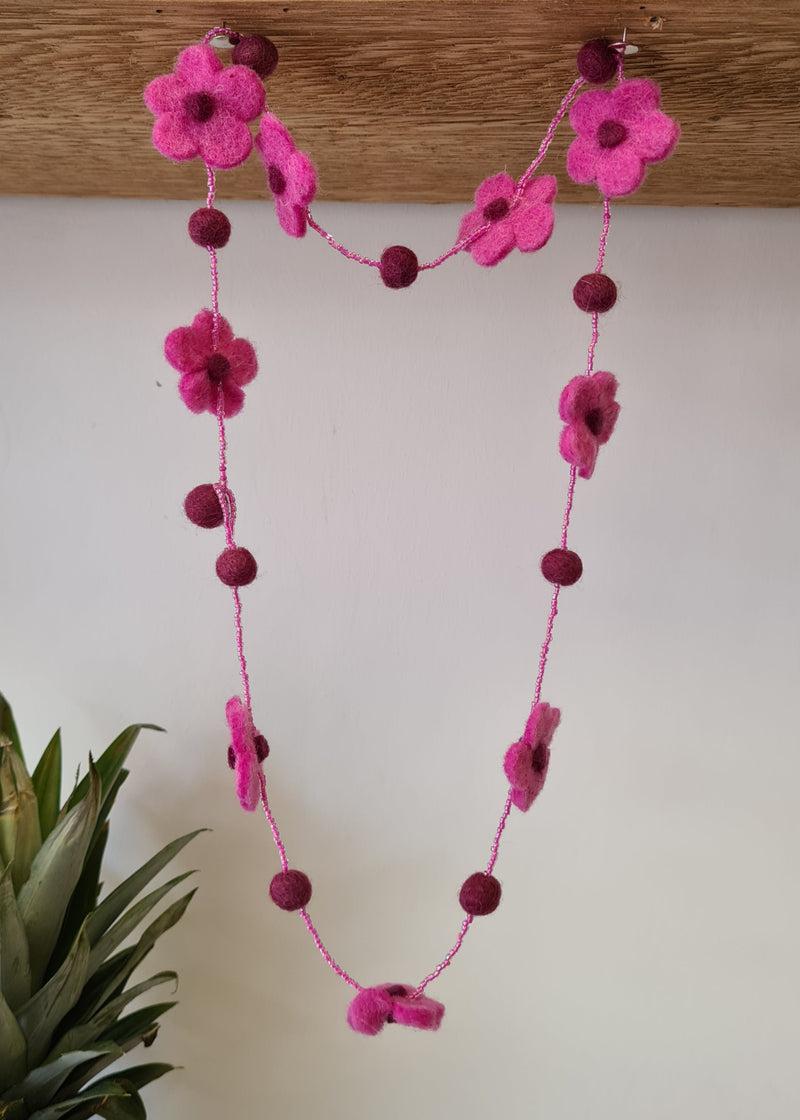 Pink pom pom flower necklace with beads hanging from a block of wood next to the top of a pineapple 