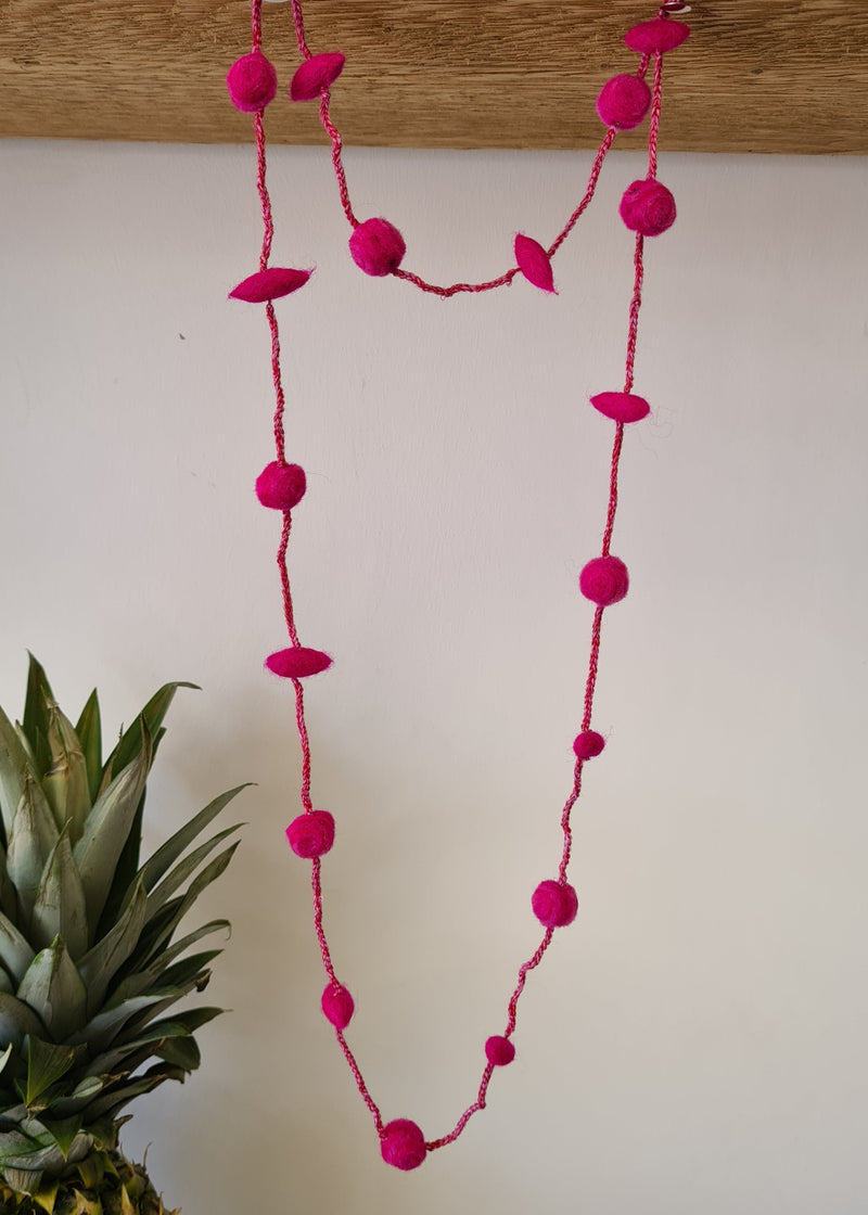 Pink Pom Pom Felt Necklace hanging from a plank of wood next to a pineapple