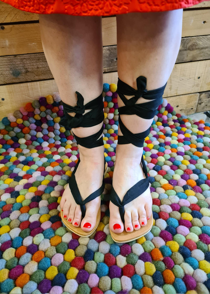 close up of woman wearing handmade black lace up sandals tied in gladiator style standing on a bright coloured pompom matt